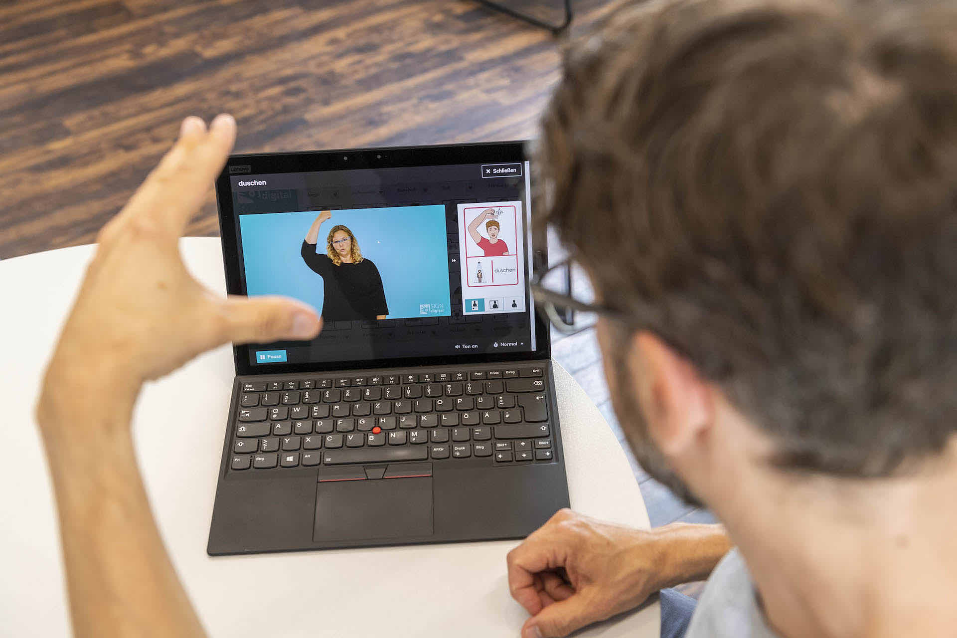 Mann gebärdet „duschen” während das SIGN-Video der Gebärde auf einem Laptop abgespielt wird.